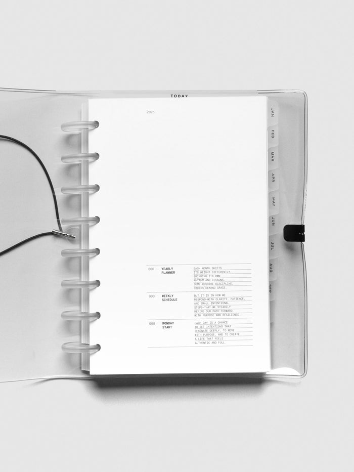 Open view of the Half Letter planner showing the first page with the 2026 title and intro text. The black monthly tab dividers from January to June are visible on the right, and the elastic closure is laid to the side.