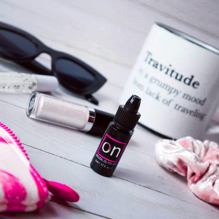 A flat-lay lifestyle shot featuring a miniature bottle of Sensuva ON Arousal Oil standing upright on a wooden desk. A white mug reading "Travitude," sunglasses, and a pink scrunchie sit softly blurred in the background.