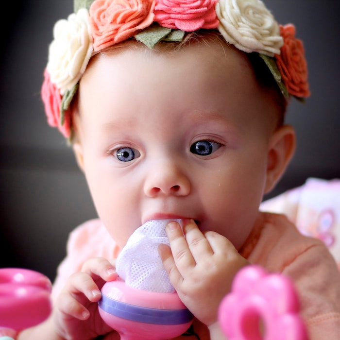 A baby in a colorful floral headband holds and chews on a Nuby The Nibbler Feeder Replacement Net (3 pack), featuring a squeeze and twist locking system, gazing forward with wide blue eyes.